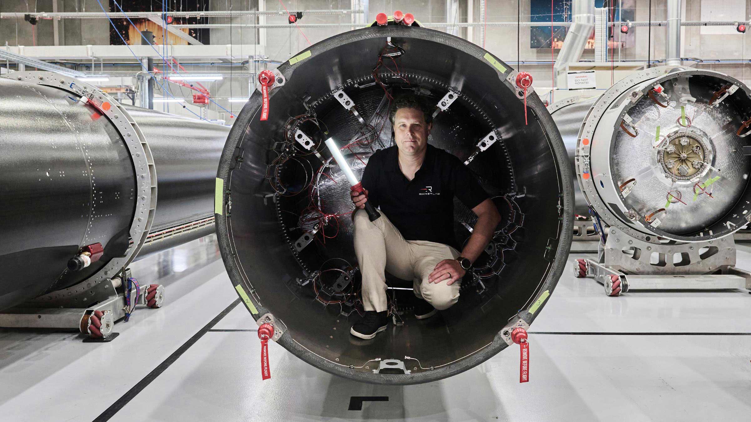 A portrait of Rocket Lab founder and CEO Peter Beck. Wearing khaki pants and a black polo shirt, Beck is crouched inside a rocket engine, holding a long flashlight.